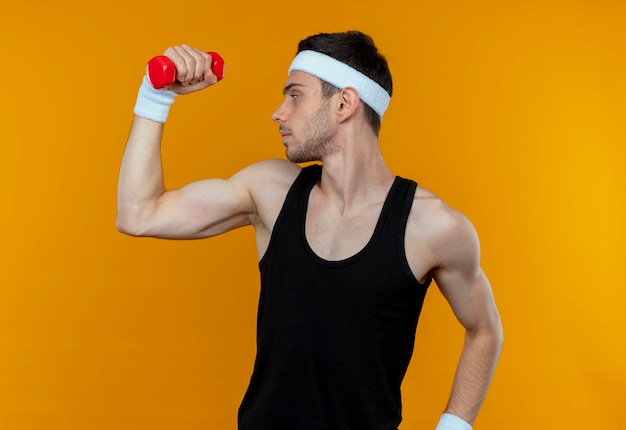 Young sporty man in headband working out with dumbbell looking confident over orange
