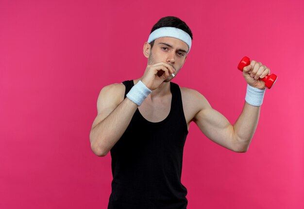 Young sporty man in headband working out with dumbbell and smoking standing over pink background