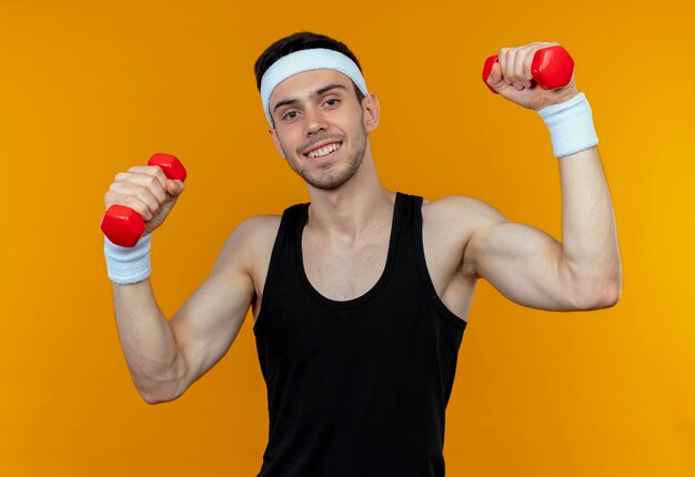 Young sporty man in headband working out with dumbbells looking at camera smiling standing over orange background