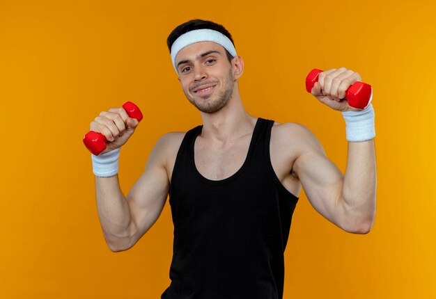 Young sporty man in headband working out with dumbbells looking at camera smiling standing over orange background