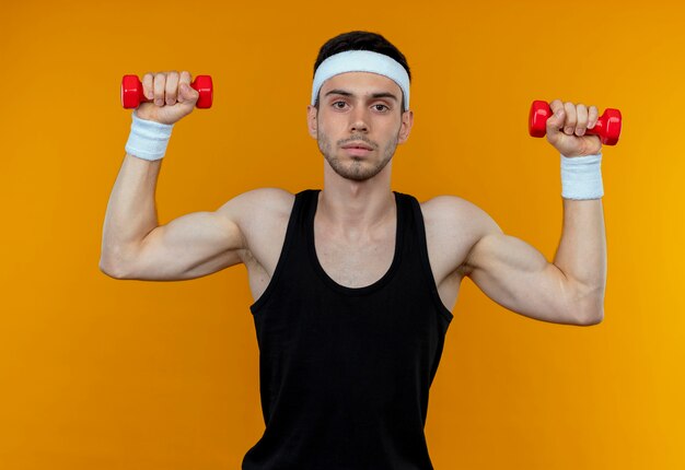 Young sporty man in headband working out with dumbbells looking at camera with serious face standing over orange background