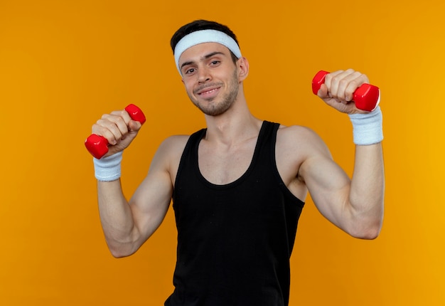 Молодой спортивный человек в повязке на голову, тренирующийся с гантелями, улыбаясь над оранжевым