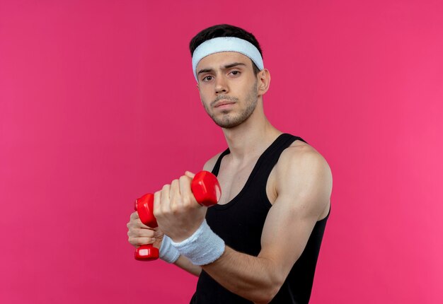 Young sporty man in headband working out with dumbbells strained and confident standing over pink wall