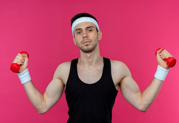 Young sporty man in headband working out with dumbbells with confident expression over pink