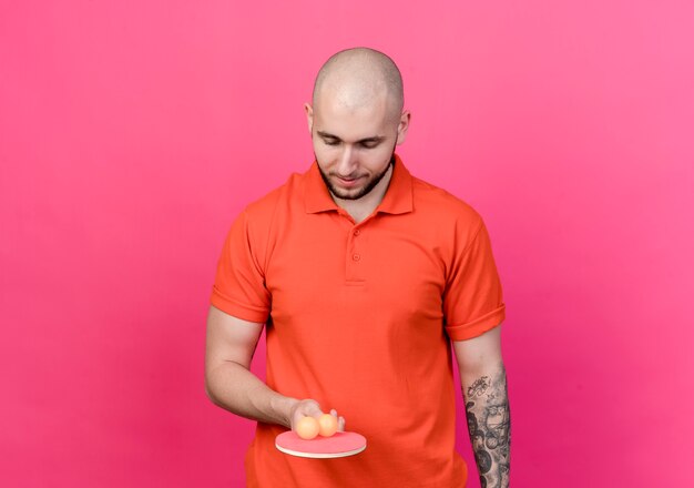 Young sporty man holding and looking at ping pong racket with balls isolated on pink wall