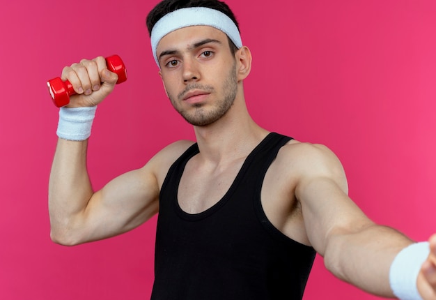 Бесплатное фото Молодой спортивный мужчина в повязке на голову, тренирующийся с гантелями, делает жест рукой над розовым