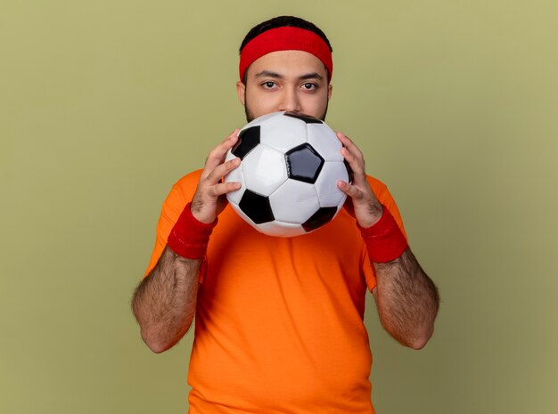 Young sporty man looking at camera wearing headband and wristband holding ball isolated on olive green