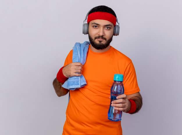 Young sporty man looking at camera wearing headband and wristband with headphones holding out water bottle at camera with towel on shoulder isolated on white background