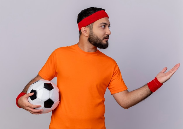 Молодой спортивный мужчина смотрит в сторону, носить повязку на голову и браслет, держа мяч и очки рукой сбоку, изолированные на белом фоне