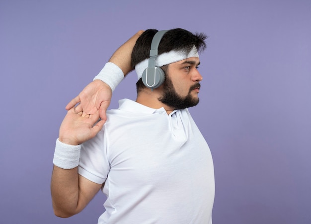 Giovane uomo sportivo in piedi in vista di profilo che indossa la fascia e il braccialetto con le cuffie che allunga il braccio