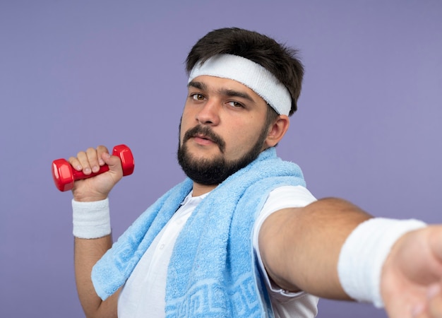 Young sporty man  wearing headband and wristband exercising with dumbbell with towel on shoulder isolated on green wall