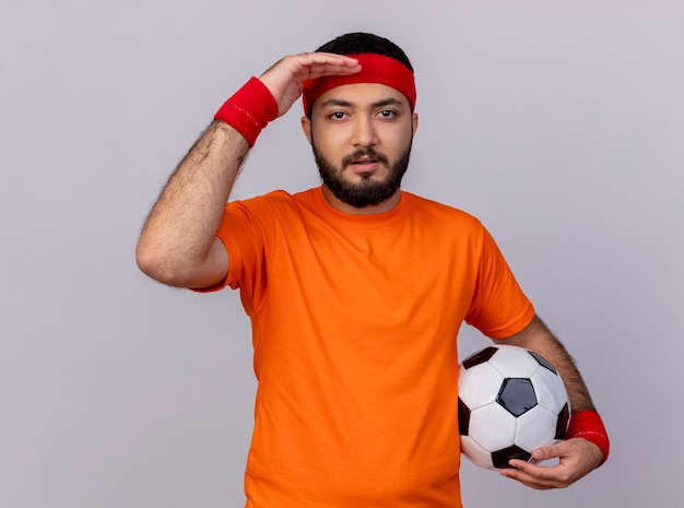 Молодой спортивный мужчина в повязке на голову и браслет, глядя в камеру с рукой, держащей мяч на белом фоне