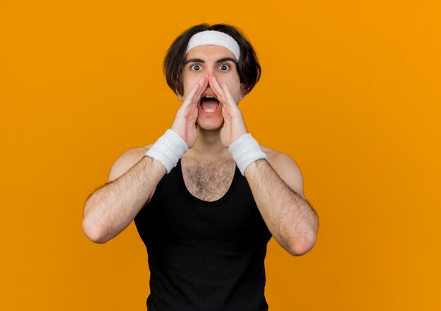 Young sporty man wearing sportswear and headband calling or shouting with hands near mouth emotional 