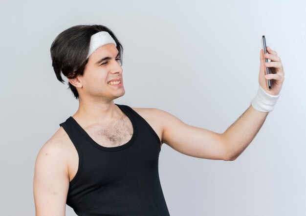 Молодой спортивный мужчина в спортивной одежде и повязке на голову делает селфи, используя свой смартфон, улыбаясь и подмигивая, стоя над белой стеной