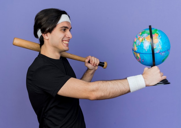 スポーツウェアとヘッドバンドを身に着けている若いスポーティな男は、元気に笑って野球のバットと地球儀を保持しています