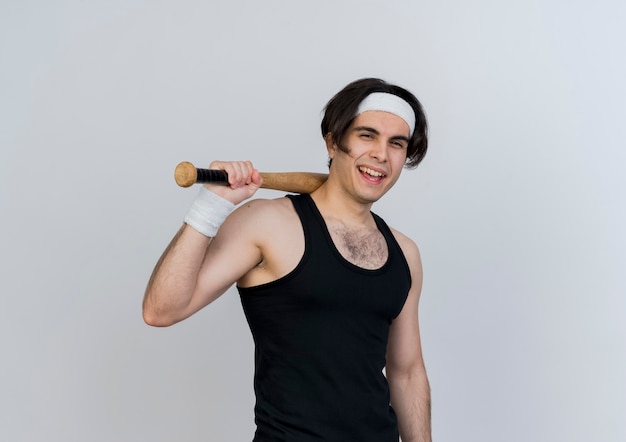 스포티 한 젊은이 입고 운동복과 야구 방망이 들고 앞을보고 웃 고 흰 벽 위에 서 윙크