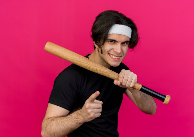 Young sporty man wearing sportswear and headband holding baseball bat pointign with index finger at camera smiling confident 