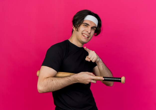Young sporty man wearing sportswear and headband holding baseball bat pointing with index finger smiling with happy face 