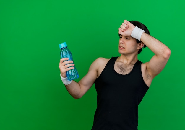 Young sporty man wearing sportswear and headband holding bottle of water looking tired 