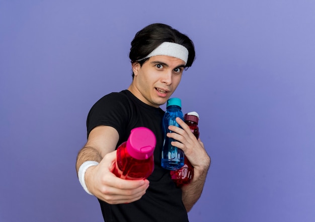 Young sporty man wearing sportswear and headband holding bottles of water offering one of bottle 