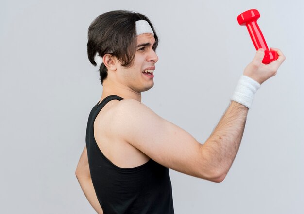 Young sporty man wearing sportswear and headband holding dumbbell looking at front confused standing over white wall