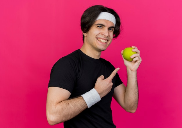 Young sporty man wearing sportswear and headband holding green apple pointing with index finger at it 