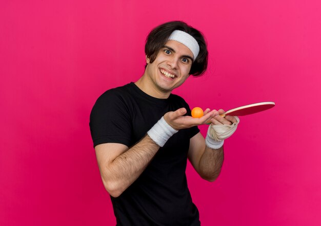 Young sporty man wearing sportswear and headband holding racket and ball for table tennis presenting with arm smiling happy and positive 