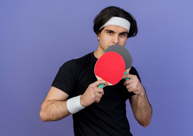 Молодой спортивный мужчина в спортивной одежде и повязке на голову с серьезным лицом держит ракетки для настольного тенниса