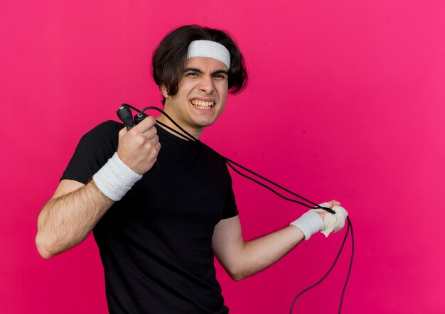 Young sporty man wearing sportswear and headband holding skipping rope displeased with annoyed expression 