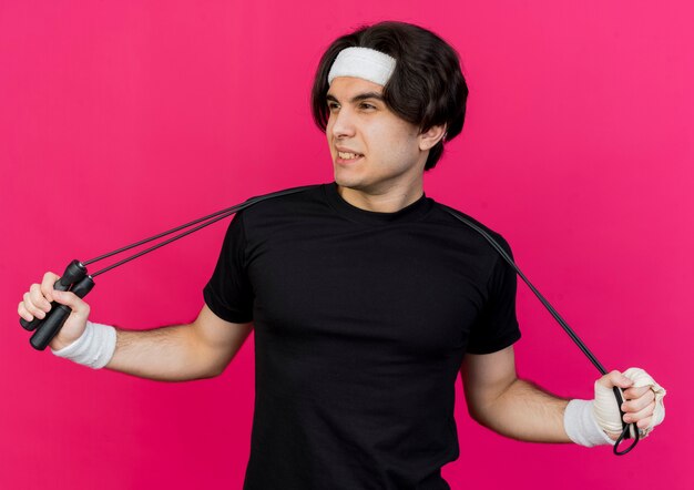 Young sporty man wearing sportswear and headband holding skipping rope looking aside smiling 