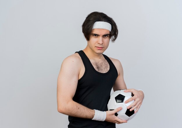 Young sporty man wearing sportswear and headband holding soccer ball looking at front with serious face standing over white wall