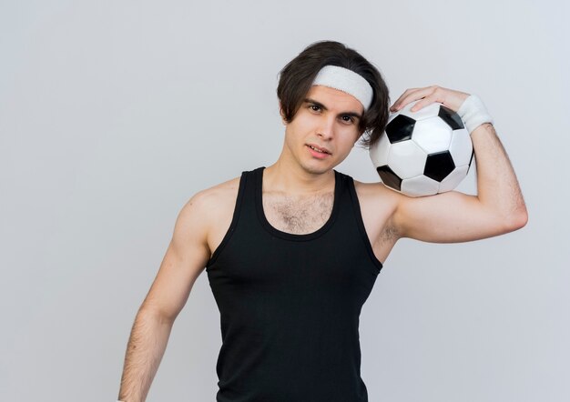 흰색 벽 위에 서 자신감 미소로 정면을보고 어깨에 축구 공을 들고 운동복과 머리띠를 착용하는 젊은 스포티 한 남자