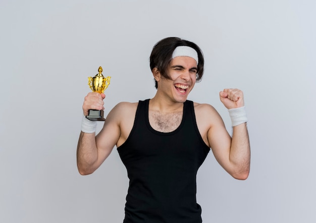 Молодой спортивный мужчина в спортивной одежде и повязке на голову держит трофей, счастливый и взволнованный, поднимающий кулак, счастливый и взволнованный, стоя над белой стеной