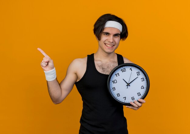Молодой спортивный мужчина в спортивной одежде и повязке на голову держит настенные часы, указывая указательным пальцем в сторону, улыбаясь стоя