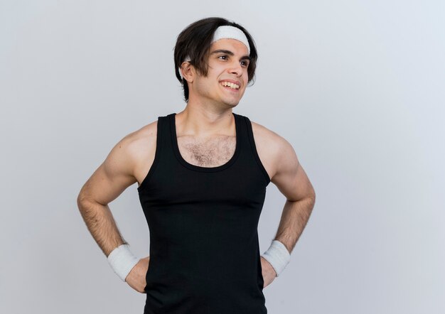 Young sporty man wearing sportswear and headband looking aside with smile on face standing over white wall