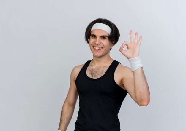 Young sporty man wearing sportswear and headband looking at front showing ok sign standing over white wall