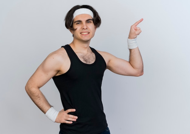 Young sporty man wearing sportswear and headband looking at front smiling confident pointing with index finger to the side standing over white wall