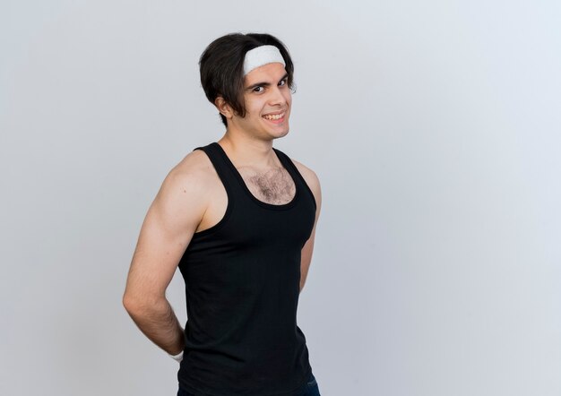 Young sporty man wearing sportswear and headband looking at front smiling slyly standing over white wall