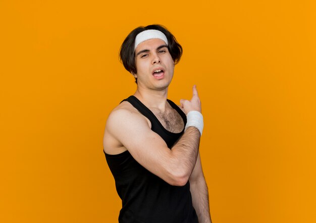 Young sporty man wearing sportswear and headband looking at front with confident expression pointing back standing over orange wall