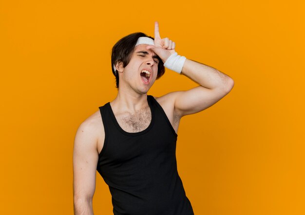 Молодой спортивный мужчина в спортивной одежде и повязке на голову делает знак проигравшего над головой и кричит с раздраженным выражением лица, стоя над оранжевой стеной