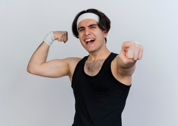 Young sporty man wearing sportswear and headband raising fist showing strength and biceps pointing with index finger atr front smiling cheerfully standing over white wall