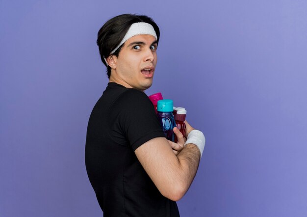 Young sporty man wearing sportswear and headband scared holding bottles of water 