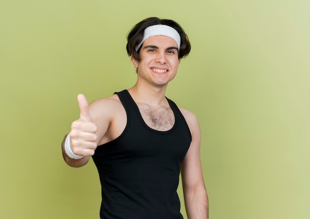 スポーツウェアとヘッドバンドを身に着けている若いスポーティな男は親指を上げて笑っている