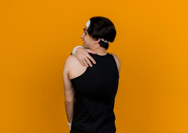 Free photo young sporty man wearing sportswear and headband standing with his back touching his shoulder over orange wall