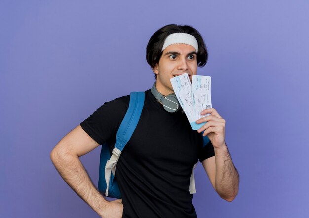 Young sporty man wearing sportswear and headband with backpack holding air tickets looking aside smiling surprised 