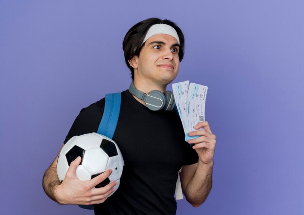 Молодой спортивный мужчина в спортивной одежде и повязке на голову с рюкзаком, держащим футбольный мяч и авиабилеты, глядя в сторону, смущенно улыбаясь