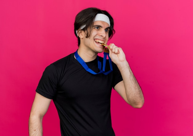 Young sporty man wearing sportswear and headband with gold medal around neck happy and excited biting medal 