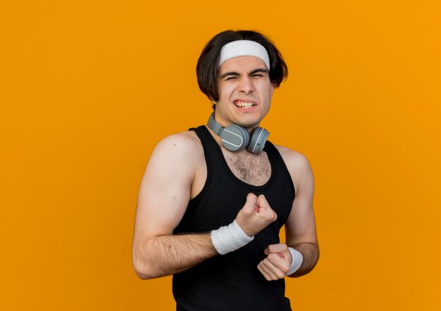 Young sporty man wearing sportswear and headband with headphones around neck looking at front clenched fists strained standing over orange wall