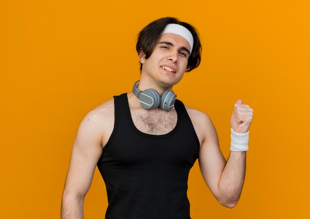 Young sporty man wearing sportswear and headband with headphones around neck looking at front smiling pointing back standing over orange wall