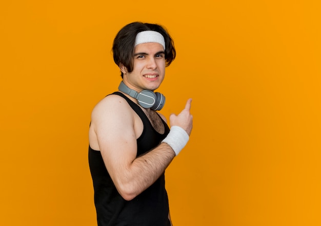 Молодой спортивный мужчина в спортивной одежде и повязке на голову с наушниками на шее, глядя вперед, улыбаясь, указывая назад, с указательным пальцем, стоящим над оранжевой стеной